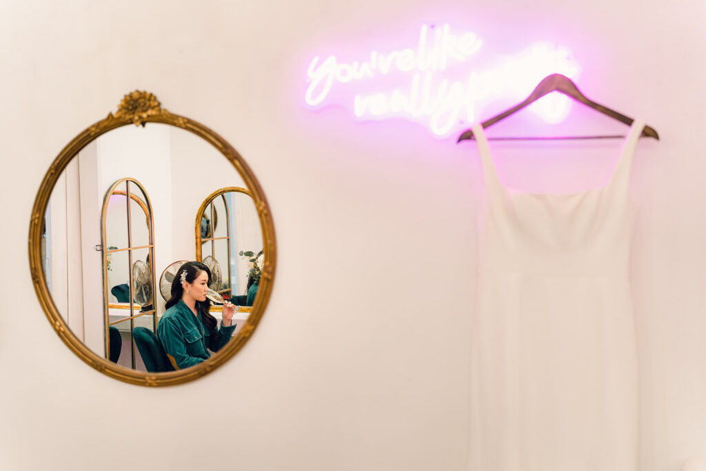 Wedding Timeline Without a Planner: Bride drinking champagne while the wedding dress hangs on the neon sign "You're like really pretty"