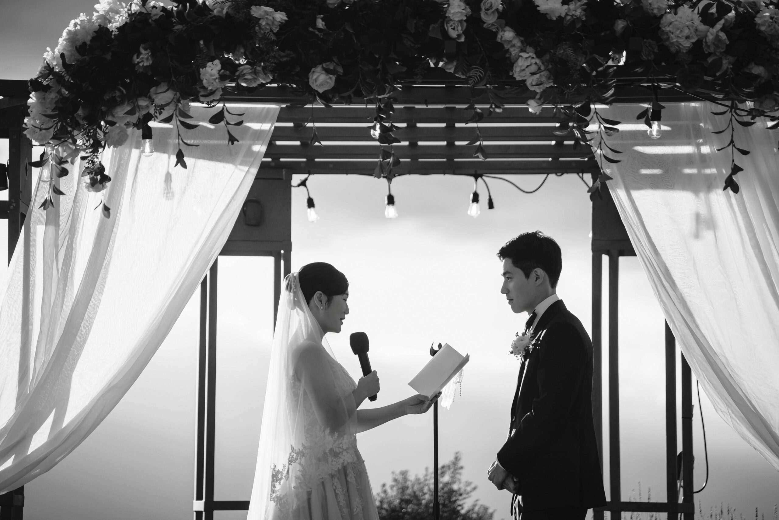 Black-and-white photograph of the bride reading her vows beneath the floral arch.