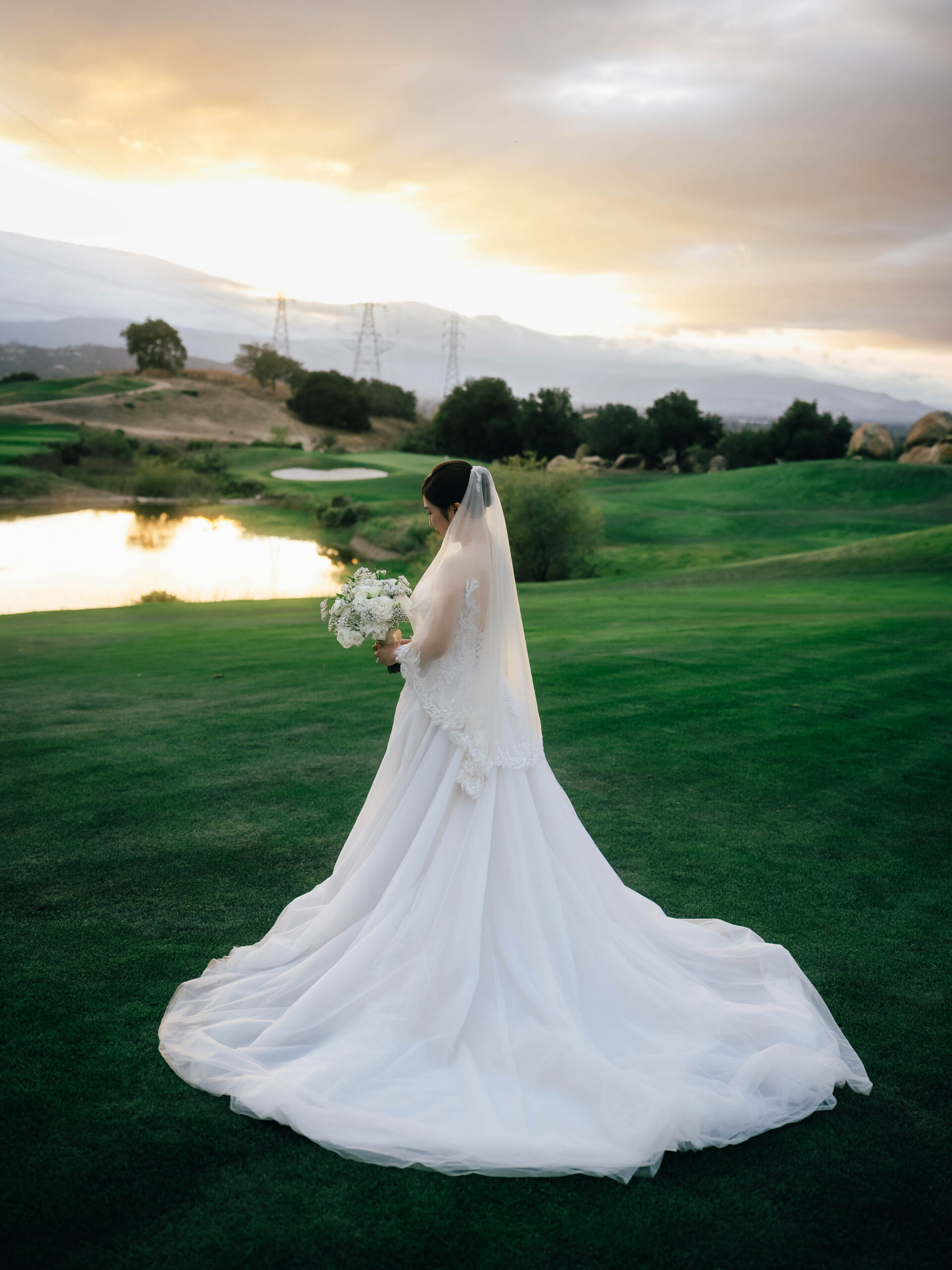 Bride holding her bouquet with her gown’s train spread across the grass beside the pond.