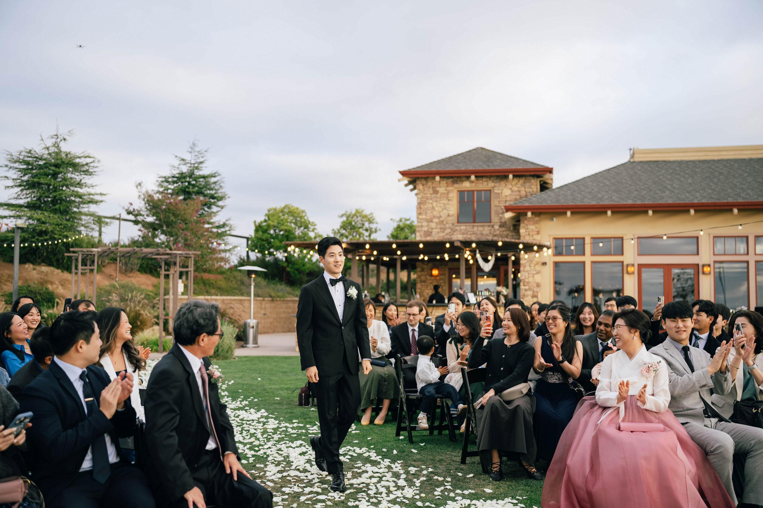Groom walking down the outdoor aisle as wedding guests applaud.