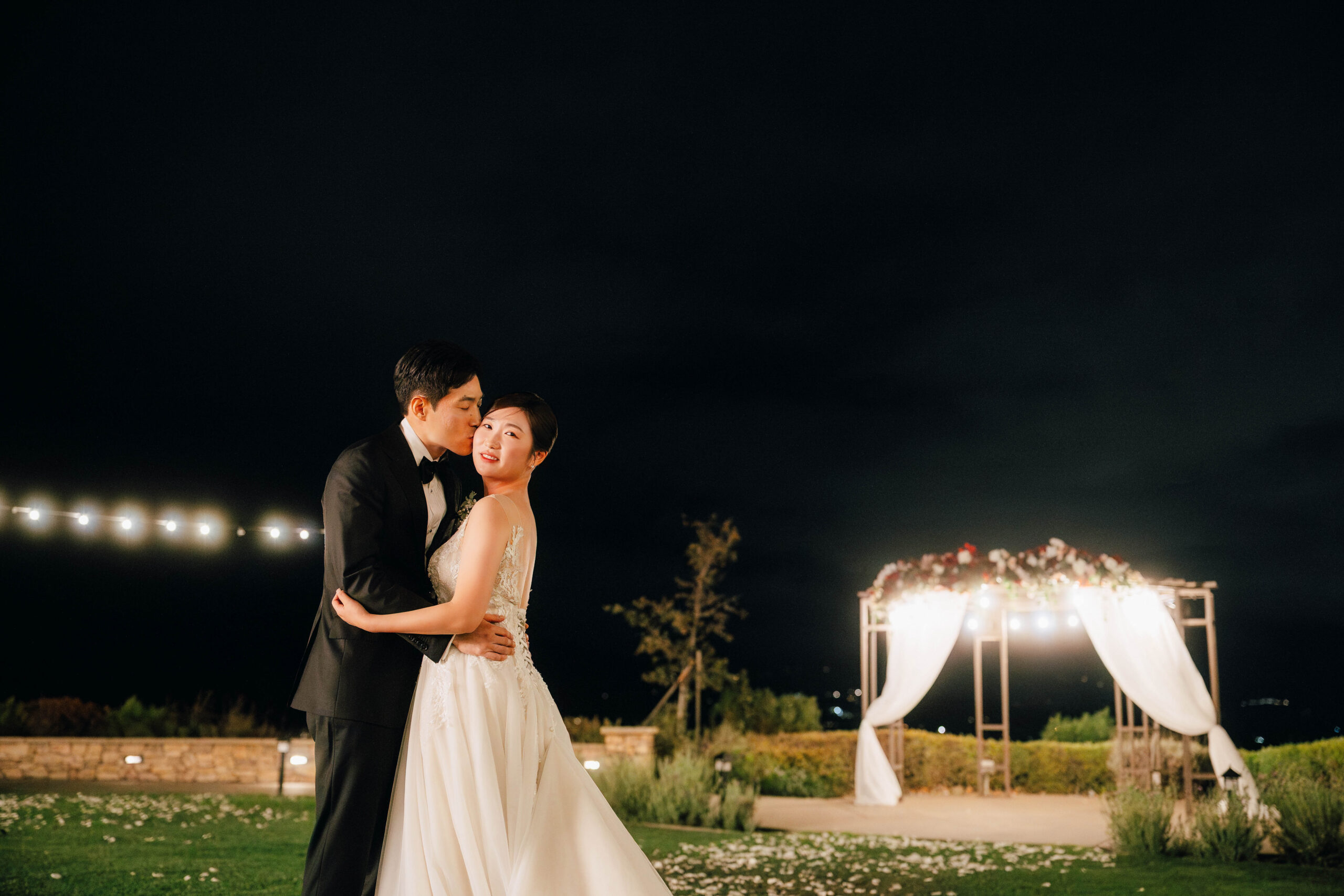 Groom kissing the bride’s cheek outside beside the illuminated ceremony arch at night.