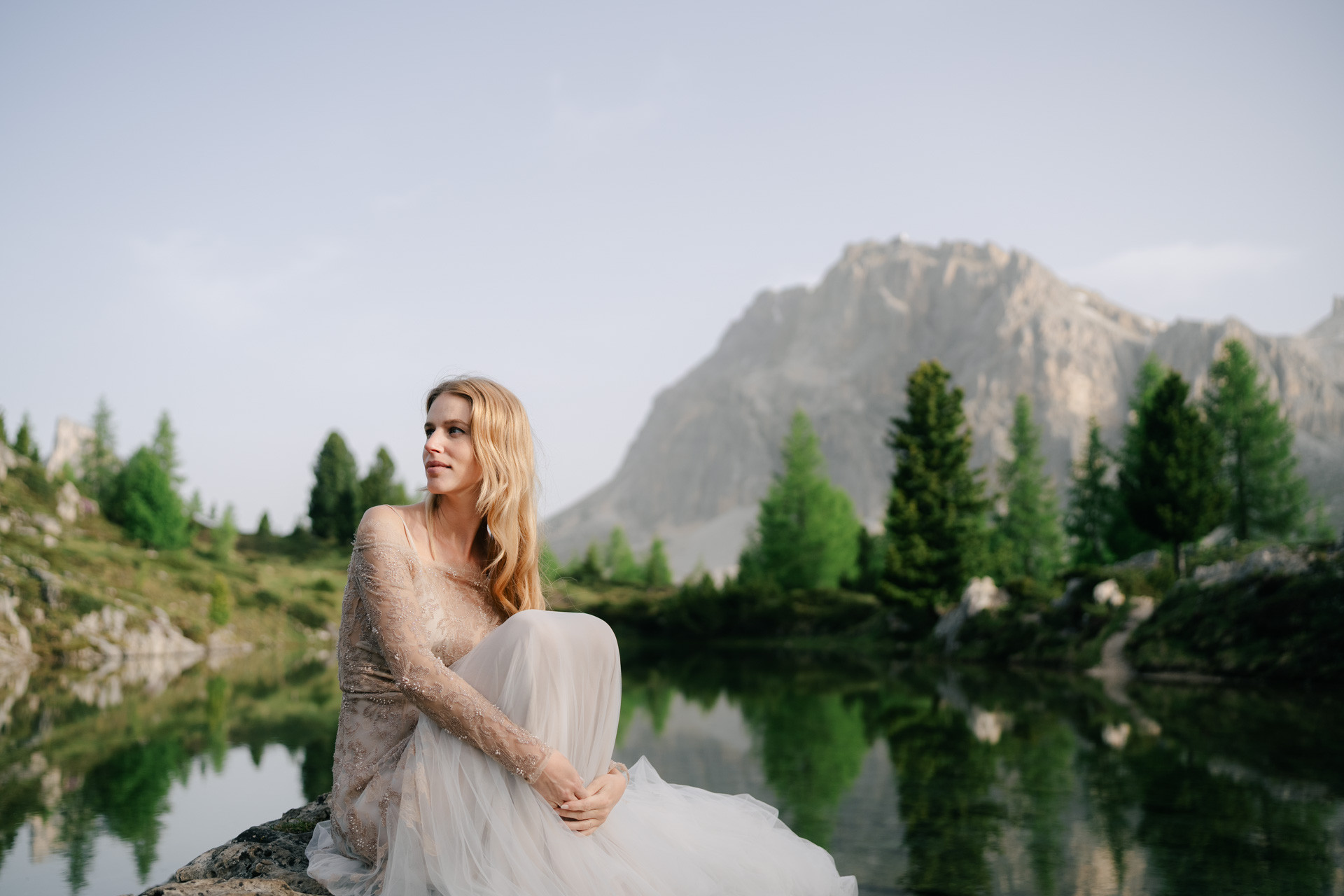 Bride seated beside a mountain lake with trees and rocky peaks behind her.