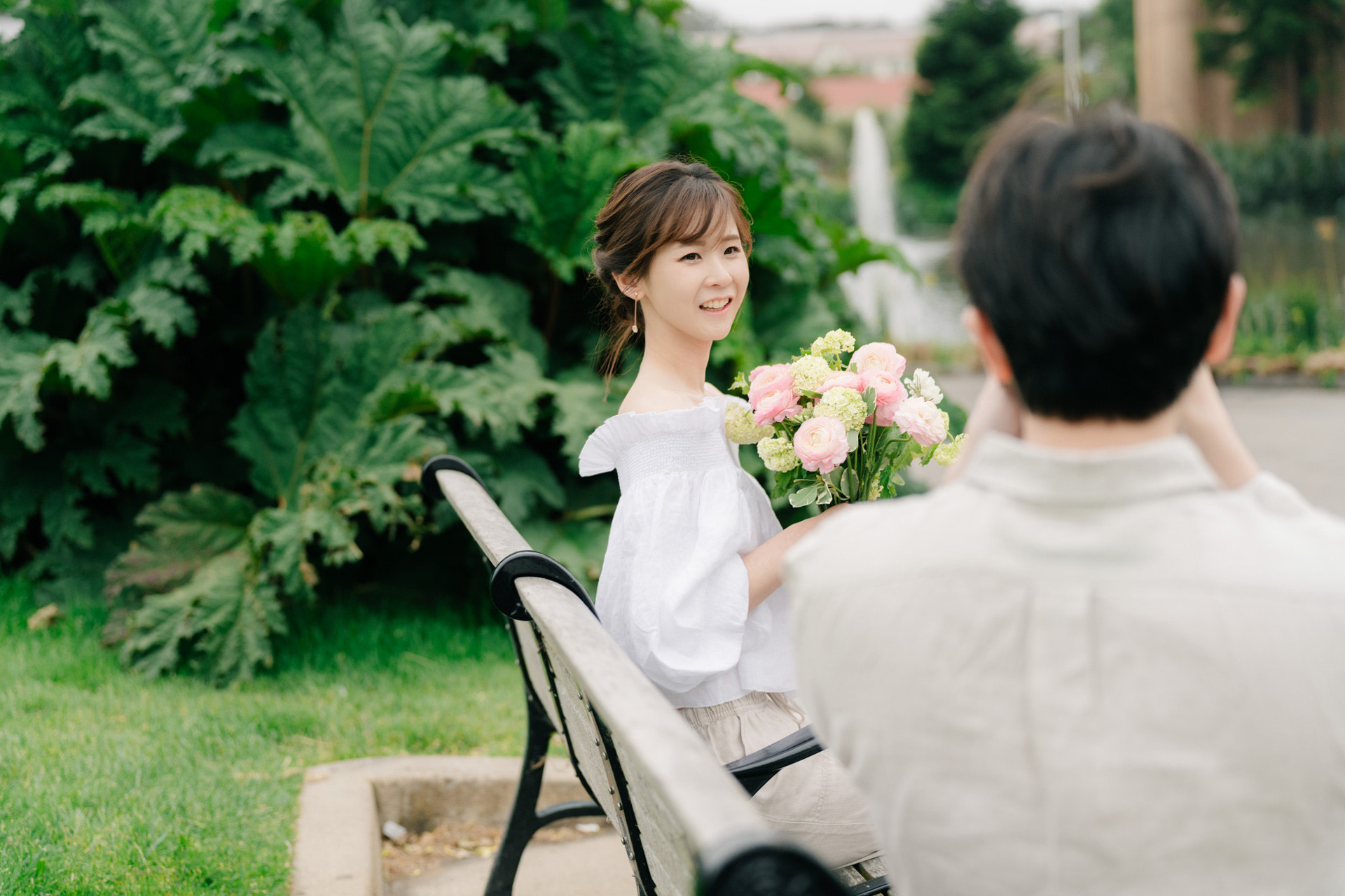 Woman smiling with a bouquet on a park bench while her partner photographs her.