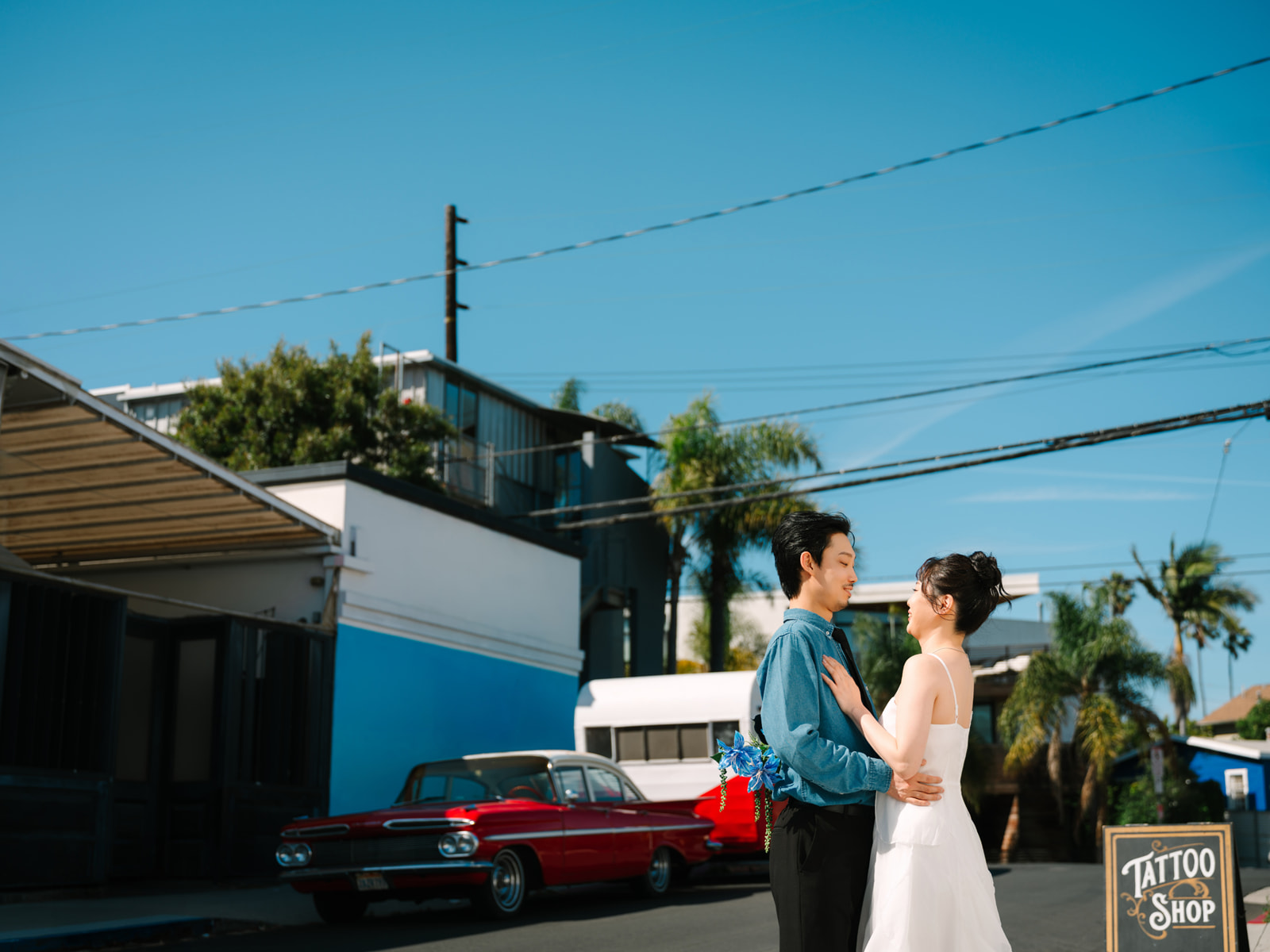 Couple embracing on a sunny street beside a red vintage car and a blue building.