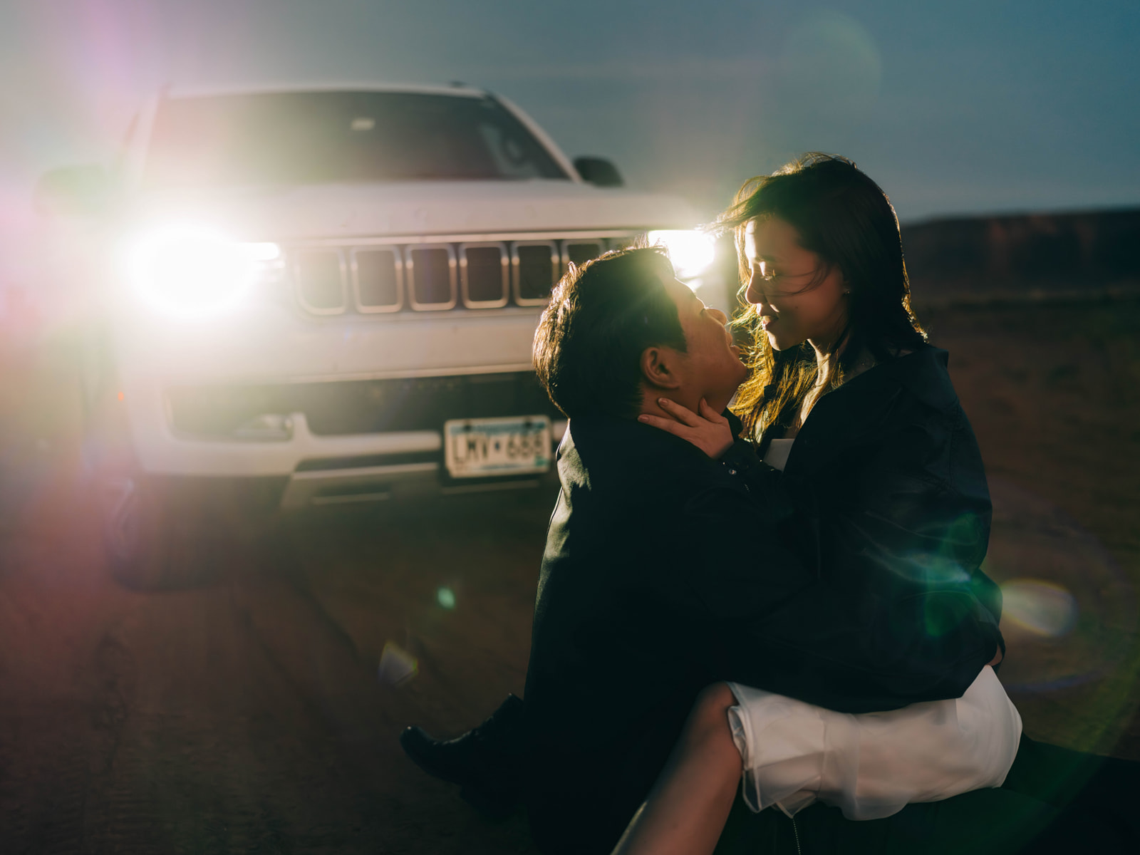 Couple embracing at dusk in the glow of vehicle headlights.