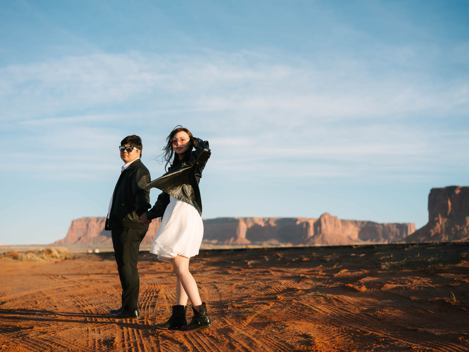 Couple in black jackets and sunglasses holding hands against a backdrop of desert mesas.