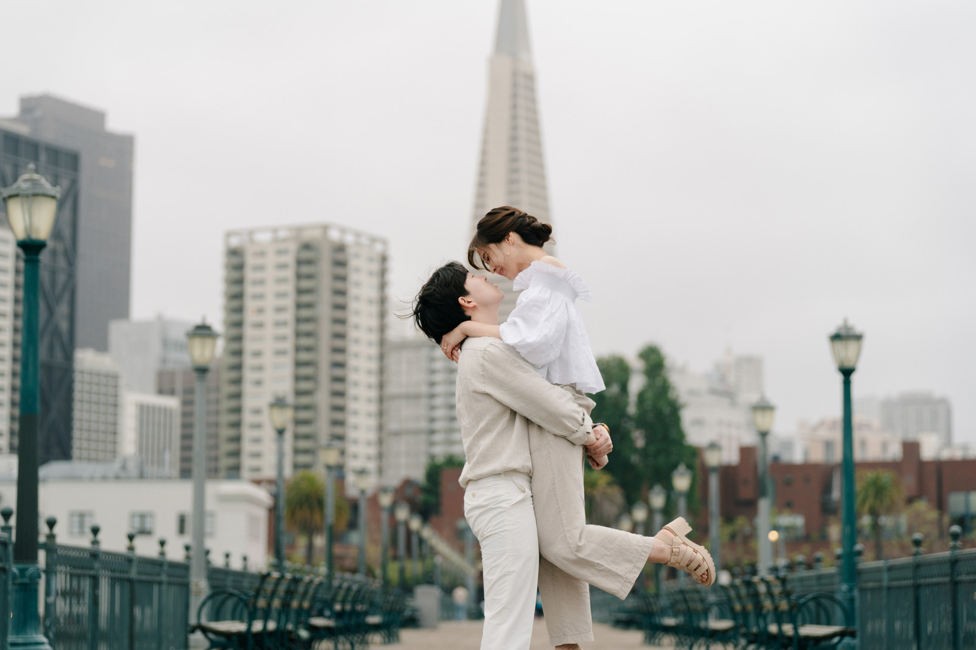Couple laughing during a lifted embrace on a pier with the San Francisco skyline behind them.
