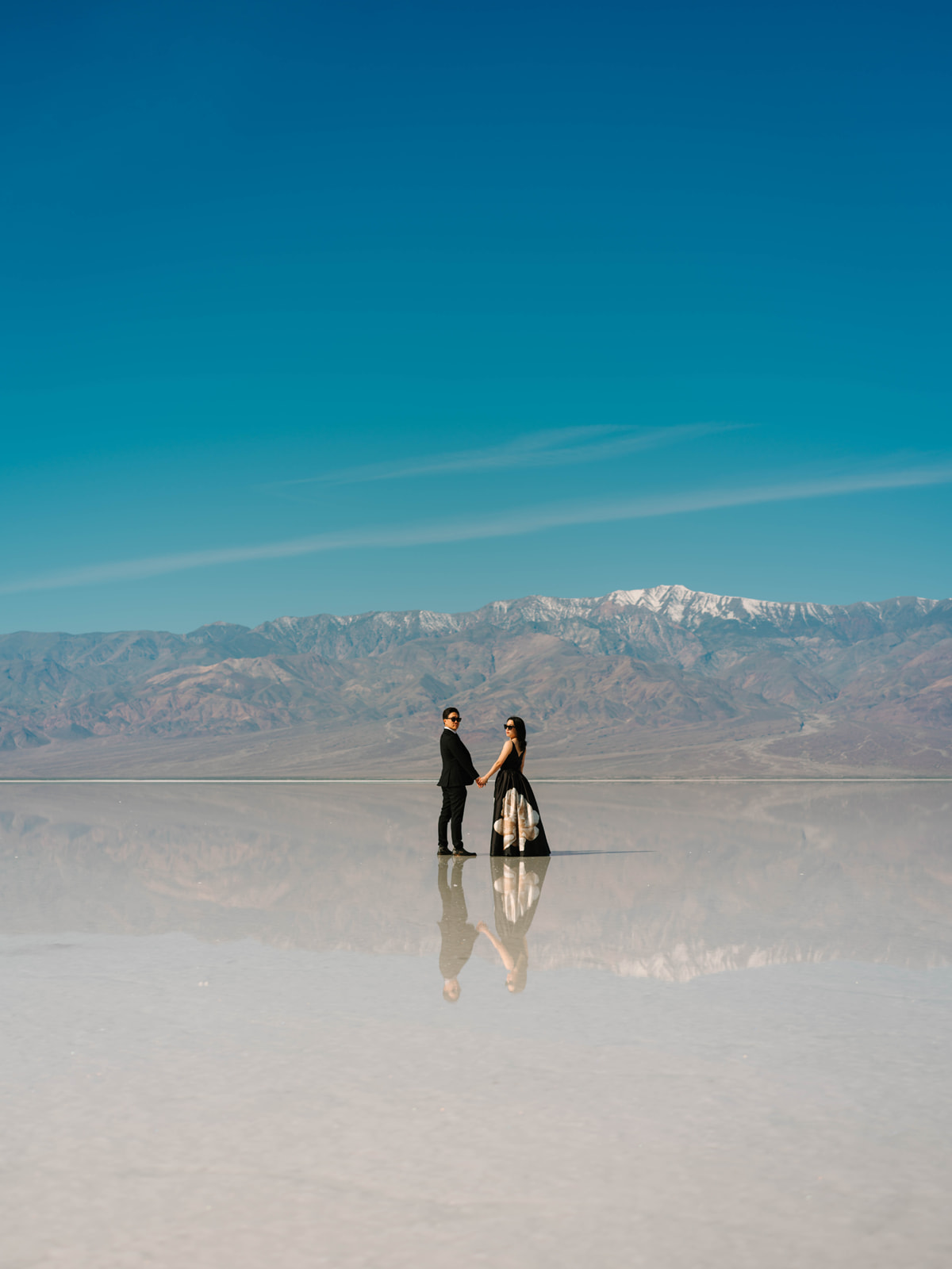 Couple holding hands on a reflective surface with mountains and blue sky behind them.