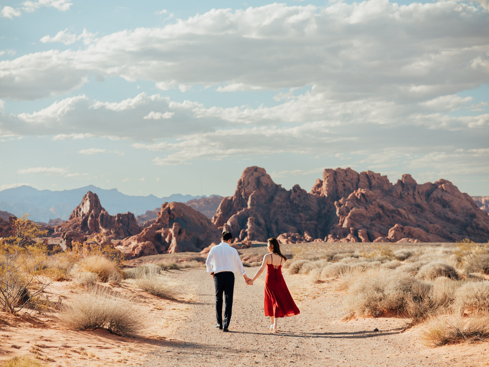 Couple holding hands as they walk toward red rock formations beneath a cloudy sky.