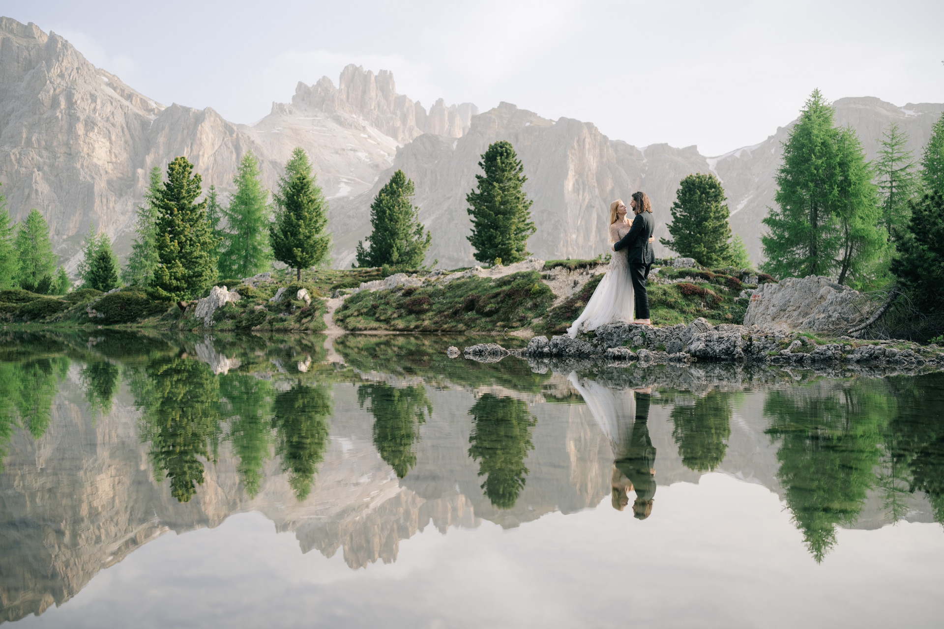 Wedding couple facing each other on rocks beside a mountain lake, reflected in the still water.
