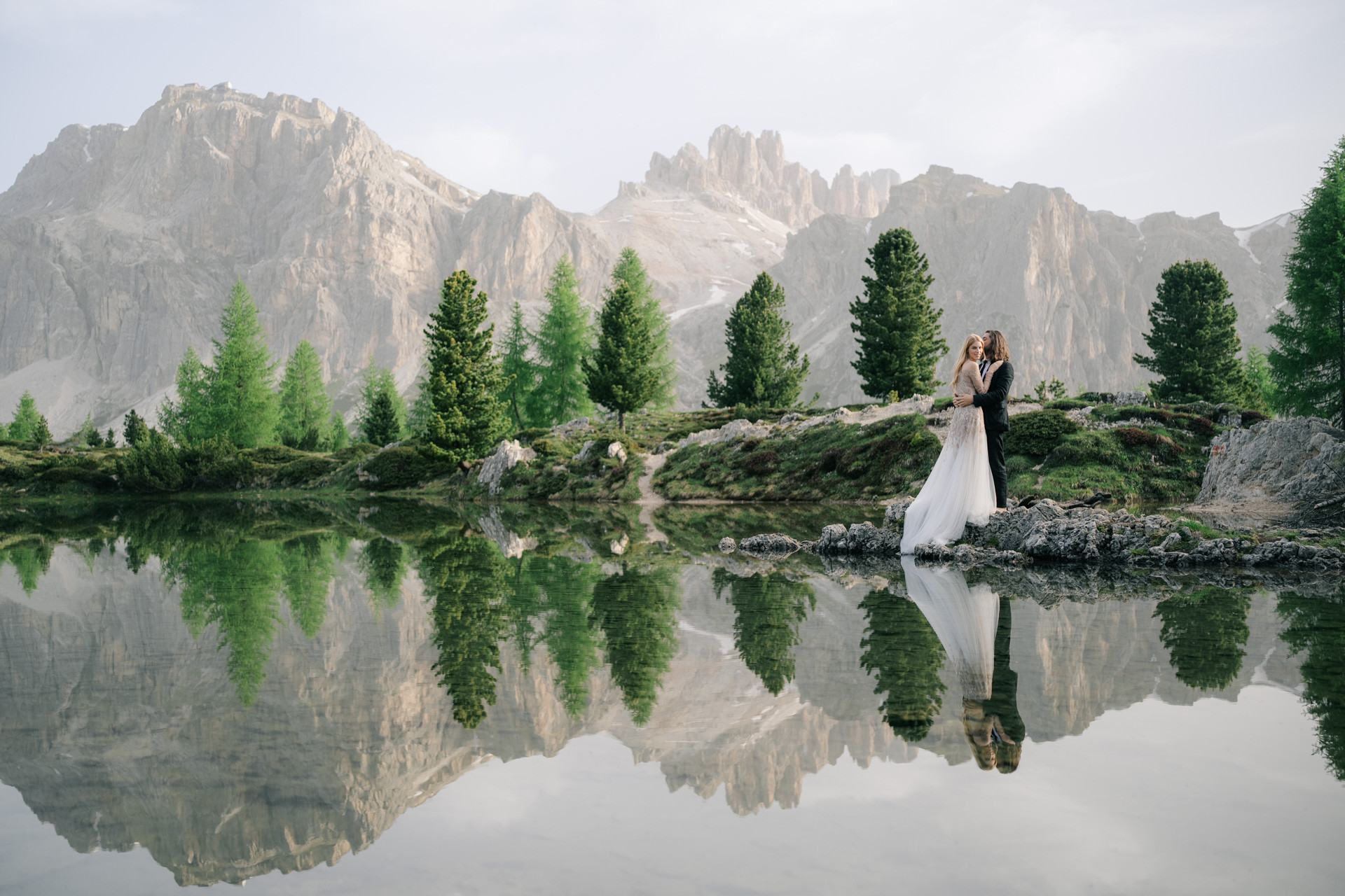 Wedding couple embracing beside a still lake reflecting trees and mountain peaks.