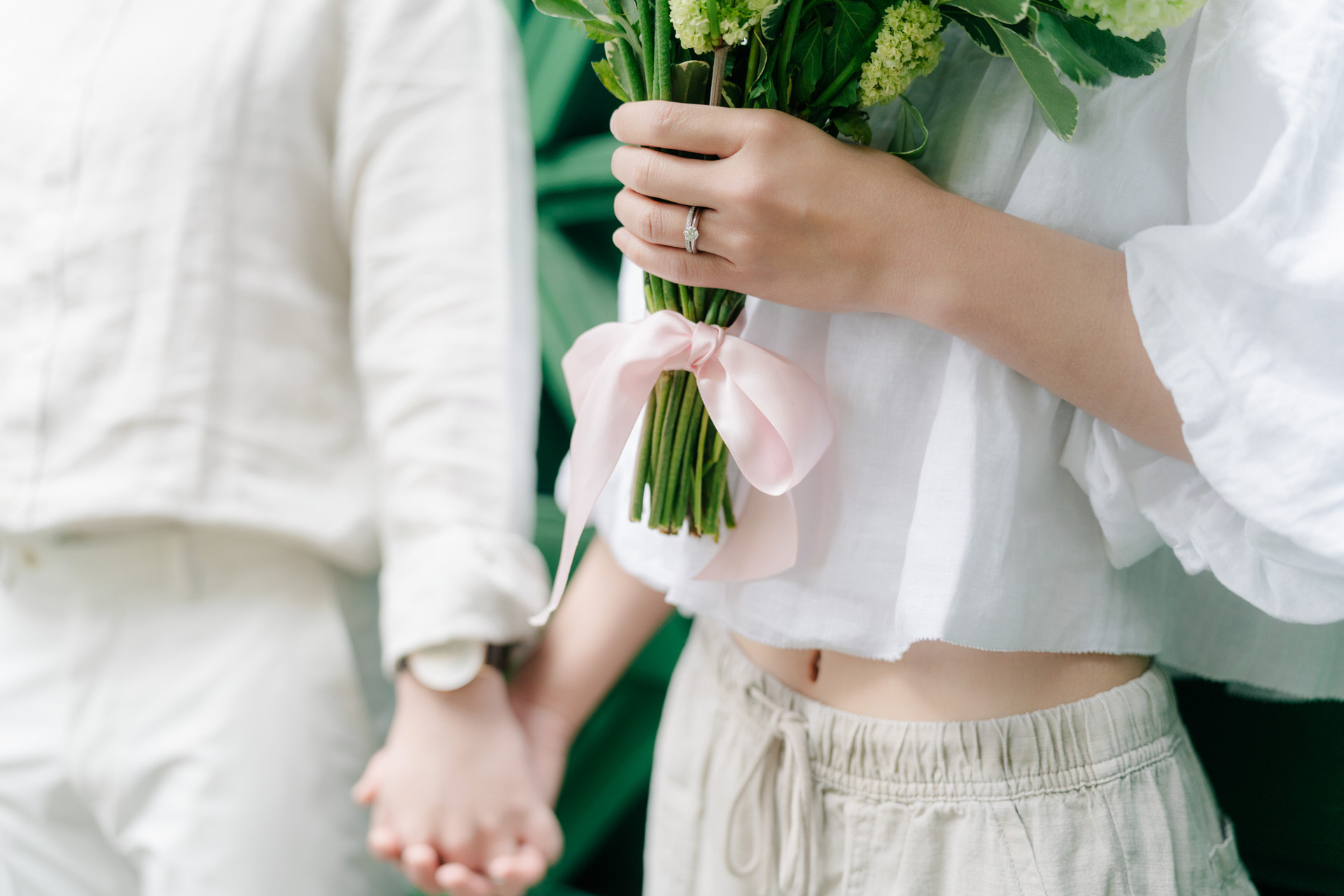 Close view of an engagement ring and pink-ribbon bouquet as a couple holds hands.