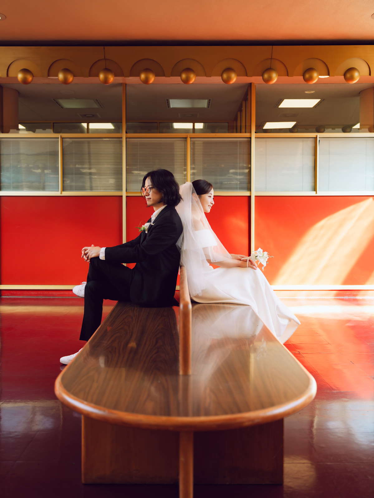 Wedding couple sitting back to back on a wooden bench in a sunlit atrium.