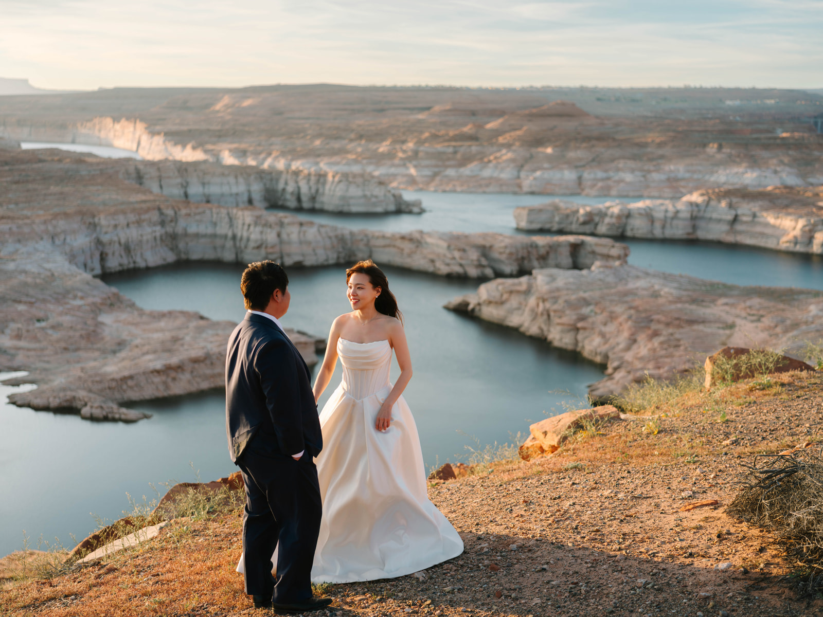 Wedding couple facing each other above winding blue water and pale canyon walls.