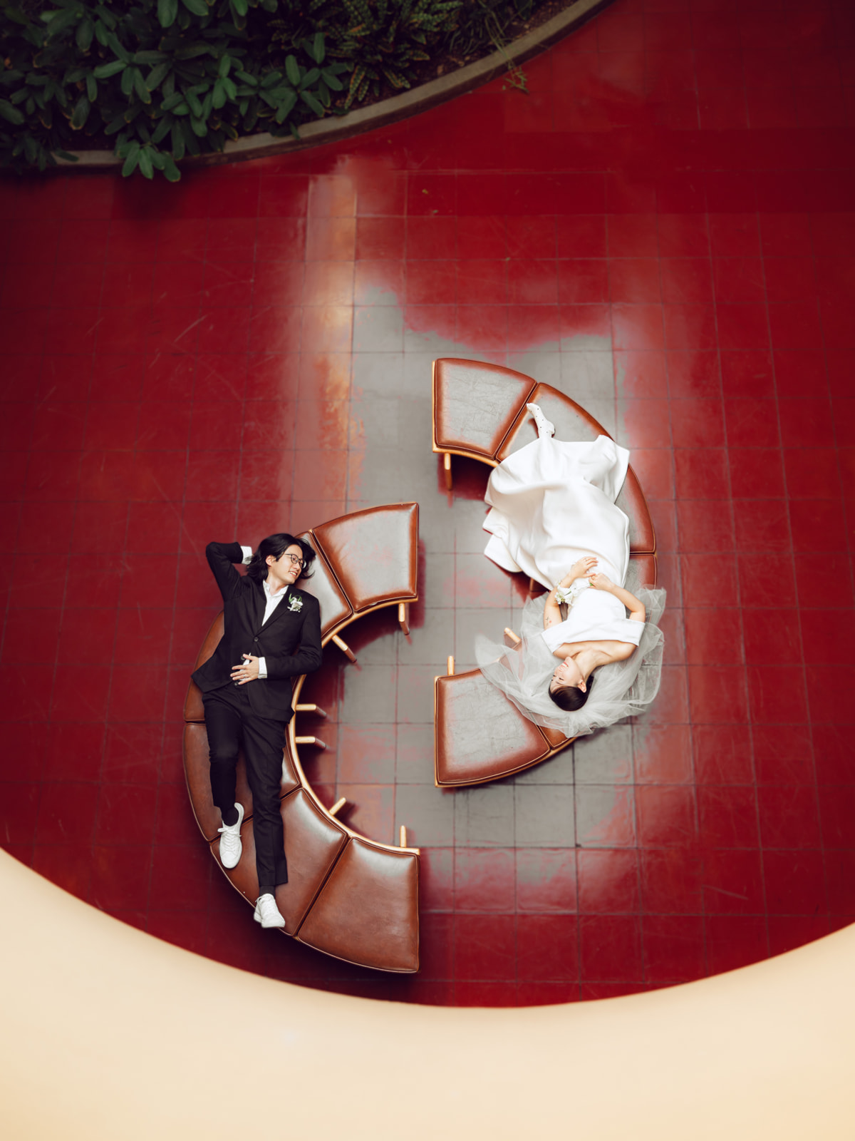 Overhead wedding portrait of a couple reclining on curved benches above red tile flooring.