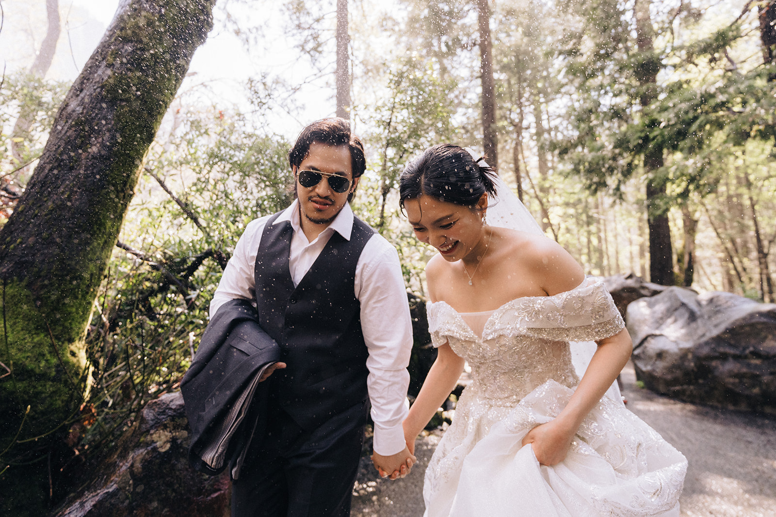 Wedding couple holding hands while walking through sunlit water spray on a forest path.