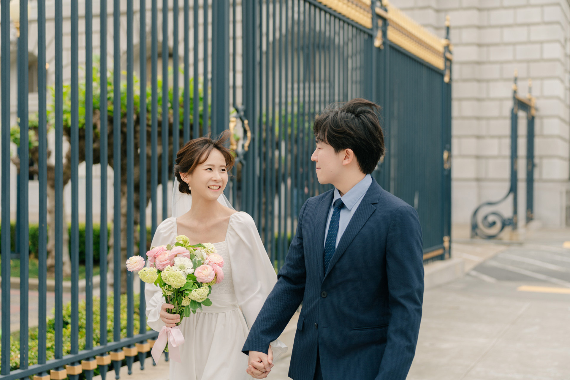 Bride and groom smiling at each other while holding hands beside a blue and gold fence.