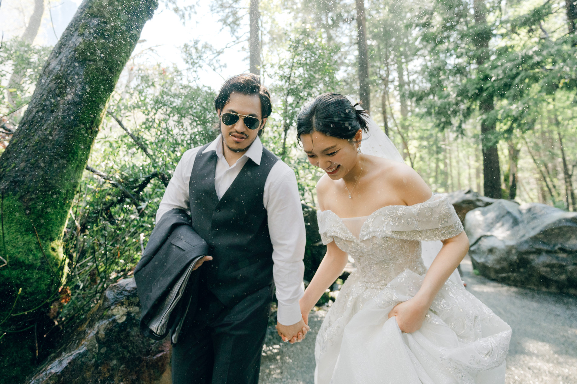 Bride and groom holding hands on a forest path surrounded by sunlit water spray.