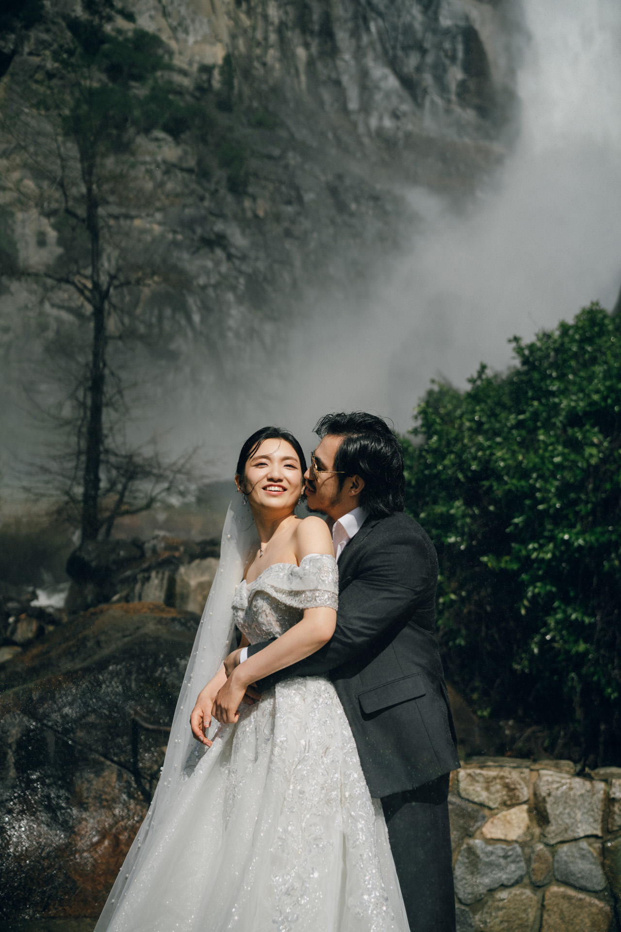 Groom kissing the bride on her cheek as they embrace in front of a waterfall.