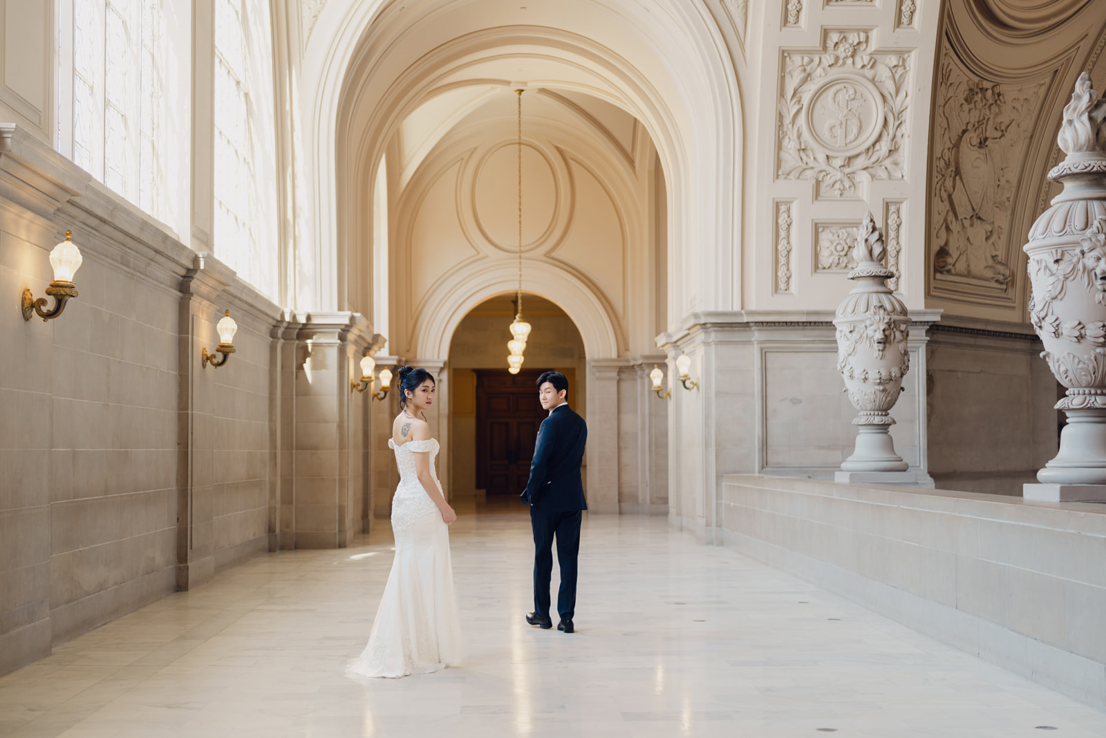 Sora in a white gown and Jonggeon in a dark suit look back in a sunlit City Hall corridor.
