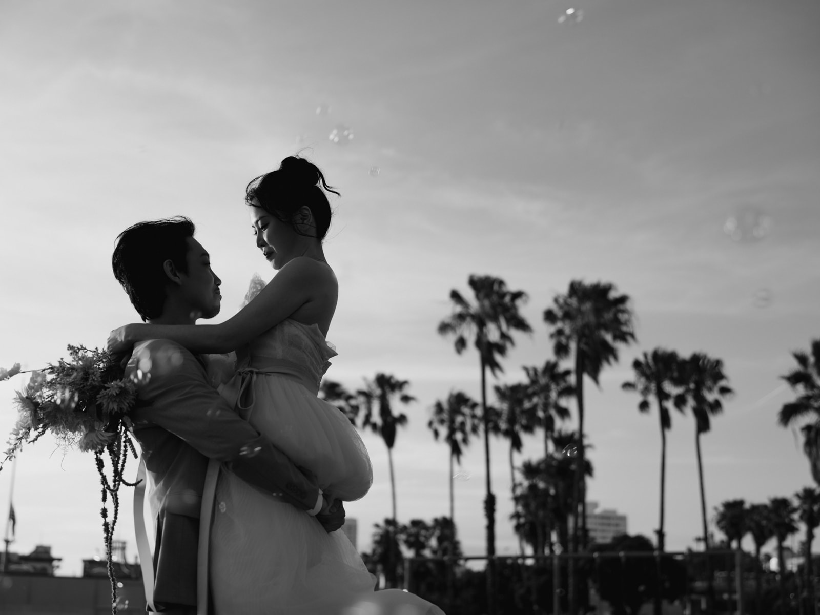 Black-and-white photograph of a lifted embrace beneath palm trees and bubbles