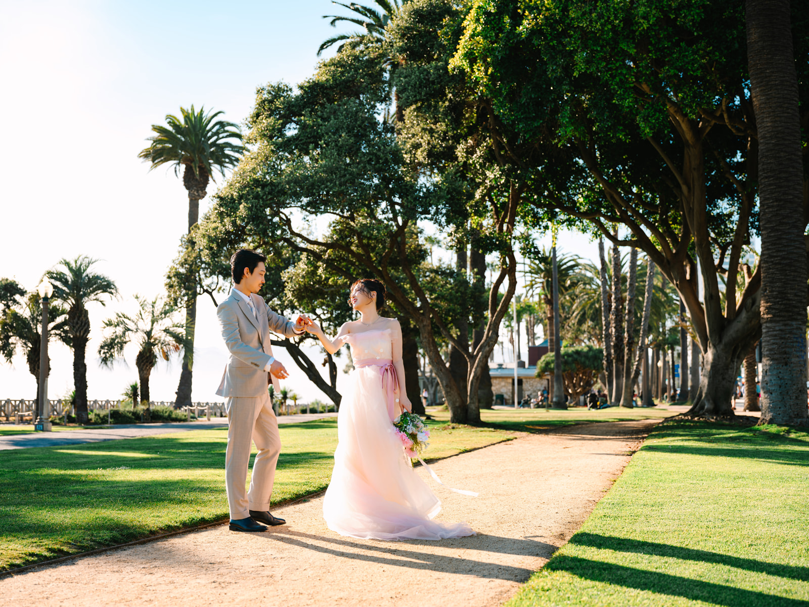 Couple twirling on a sunlit park path beneath palms and leafy trees