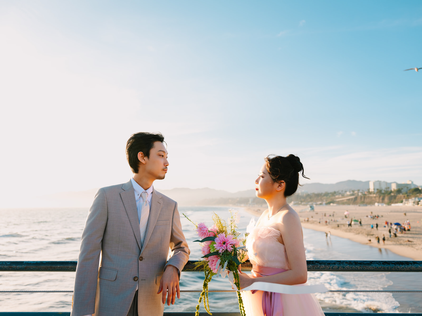 Couple looking at each other beside the pier railing with the coastline behind them