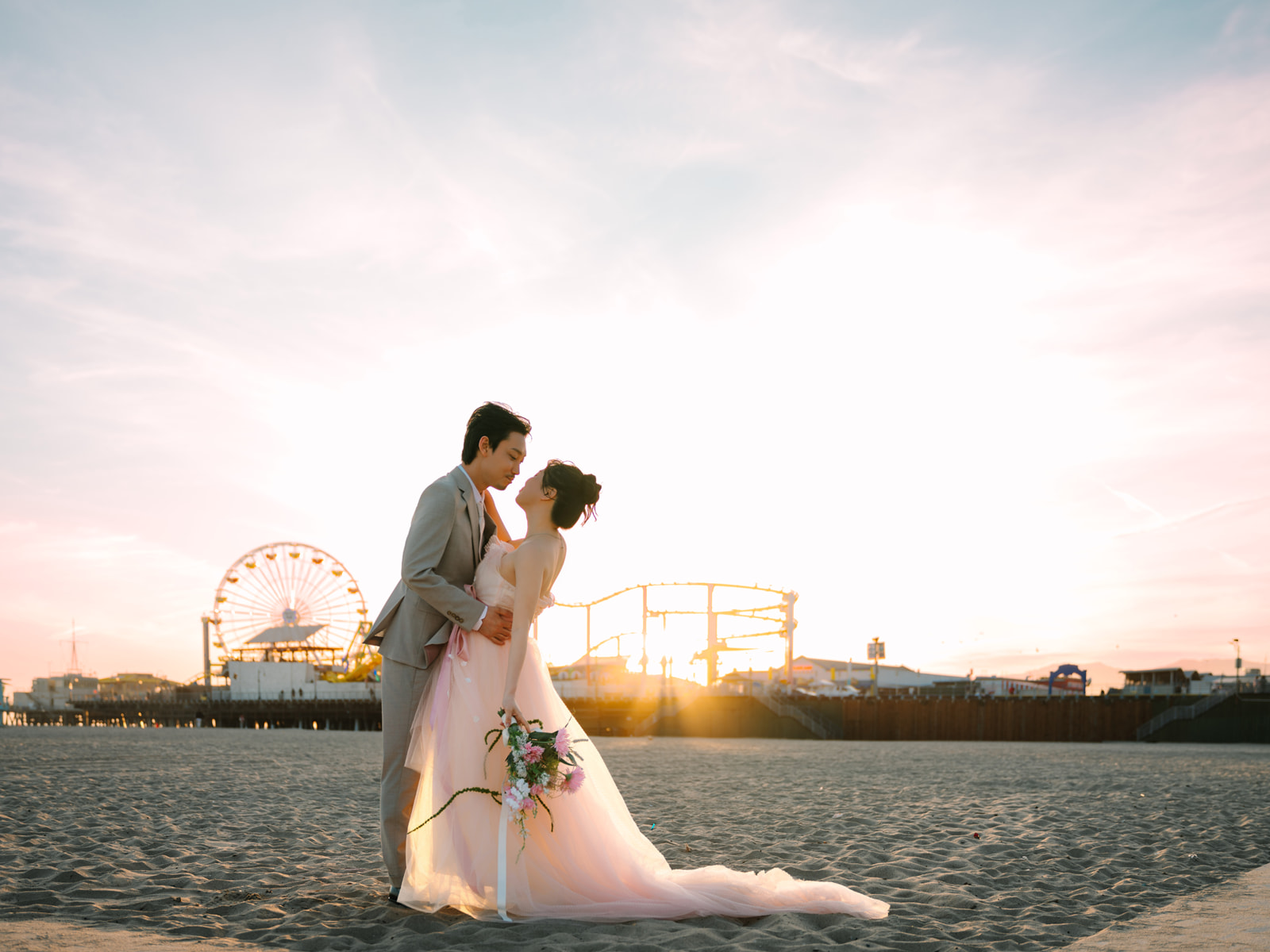 Couple leaning toward a kiss on the sand with Santa Monica Pier behind them at sunset