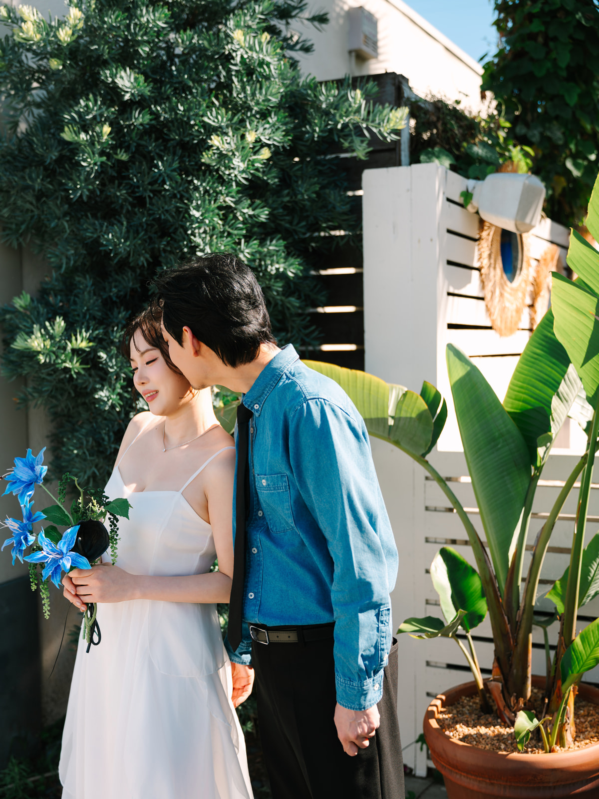 Man kissing his partner beside tropical plants and a white fence