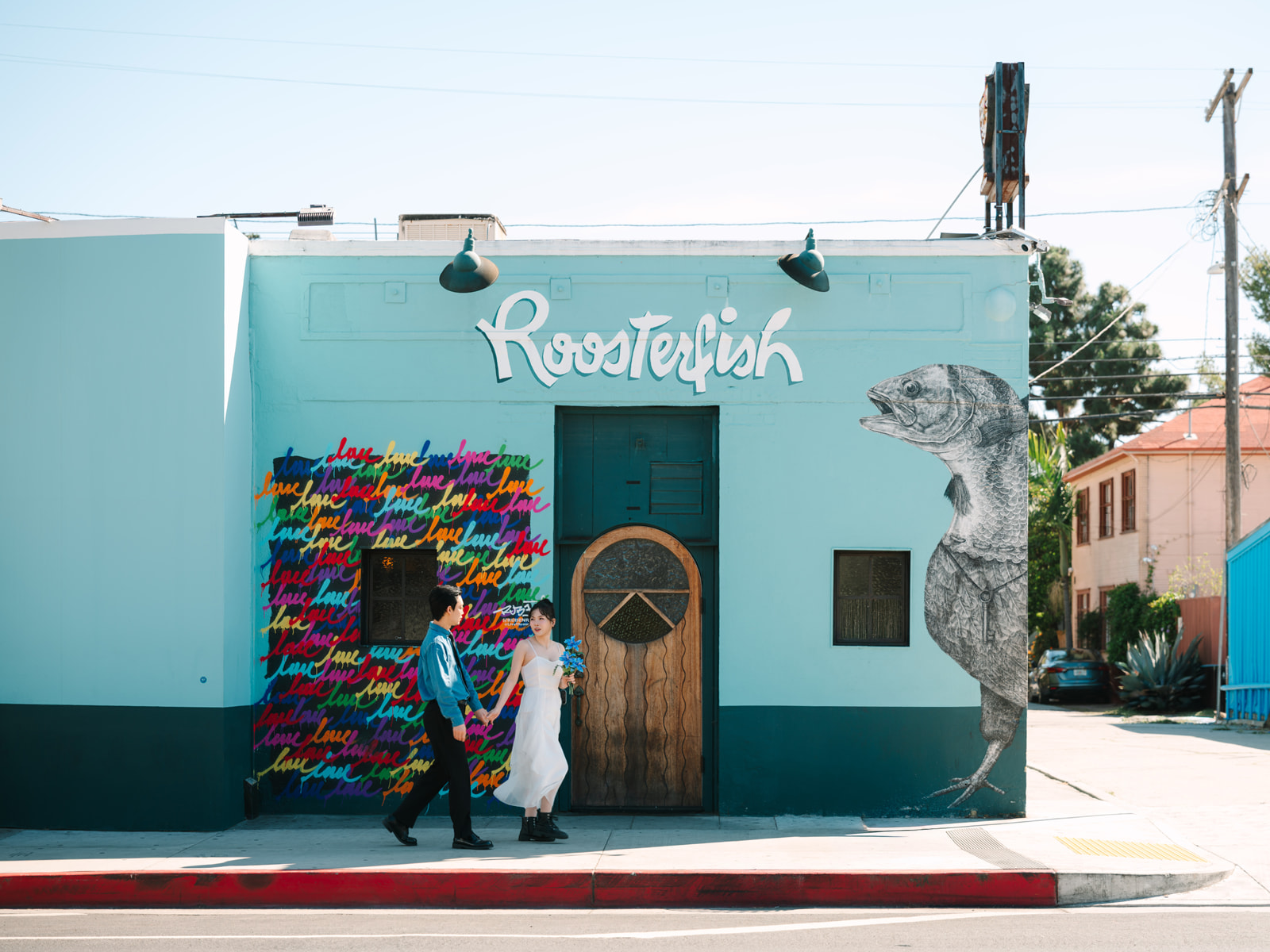 Couple walking past the turquoise Roosterfish building and colorful love mural